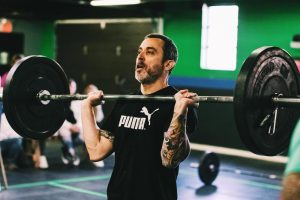A masters-age athlete holding a loaded barbell in the front rack during a Garage Games Masters Tour competition, spectators behind him in the gym