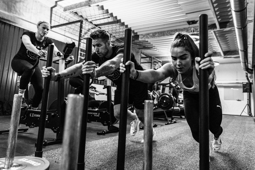 Three athletes drive weighted sleds toward the camera across an indoor race floor