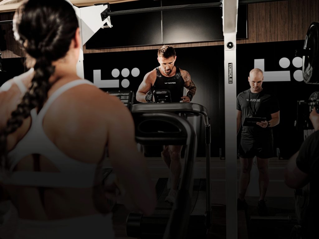 Athlete running on a treadmill during an LT Games hybrid fitness competition at Life Time, with a judge scoring alongside