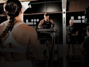 Athlete running on a treadmill during an LT Games hybrid fitness competition at Life Time, with a judge scoring alongside