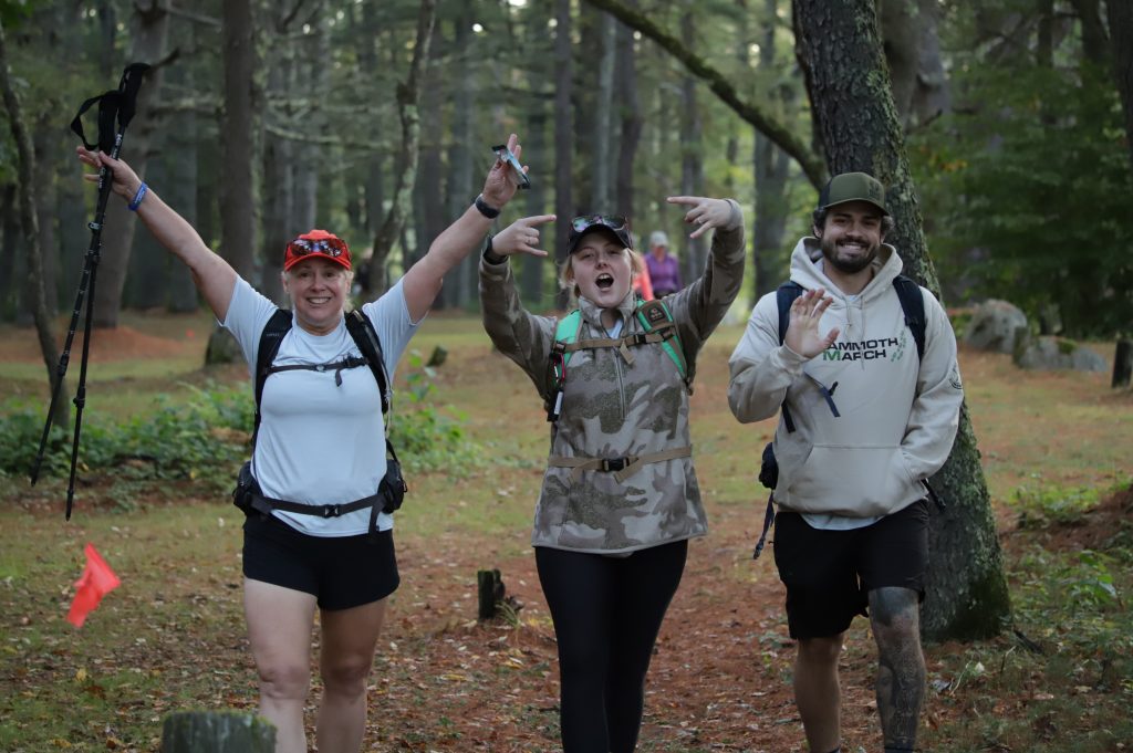 Three hikers with backpacks and trekking poles cheering on a wooded trail at Mammoth March Hudson Valley