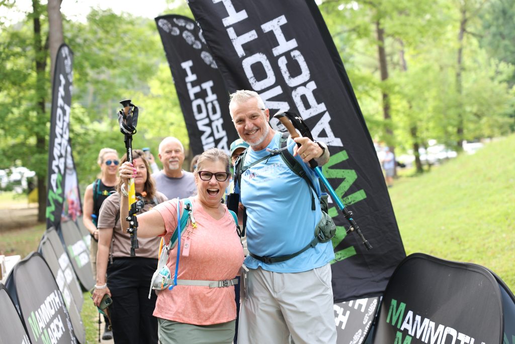 Hikers with backpacks and trekking poles beside the Mammoth March start banners in Kentucky