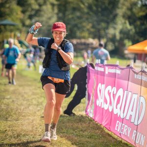 Runner passing a Sassquad Trail Running banner in a grass field at Wild Goose Trail Festival