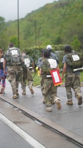 Marchers in uniform carrying rucksacks and a memorial placard along a wet mountain road at the Mountain Man Memorial March