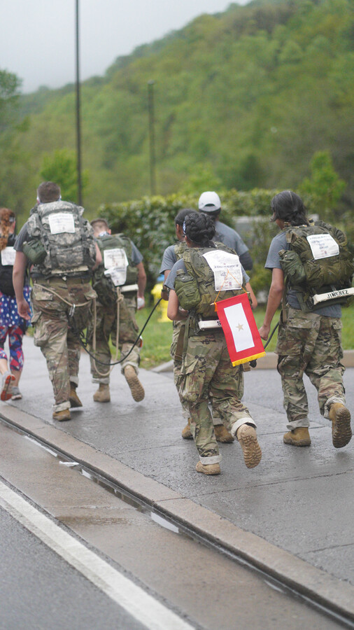Marchers in uniform carrying rucksacks and a memorial placard along a wet mountain road at the Mountain Man Memorial March