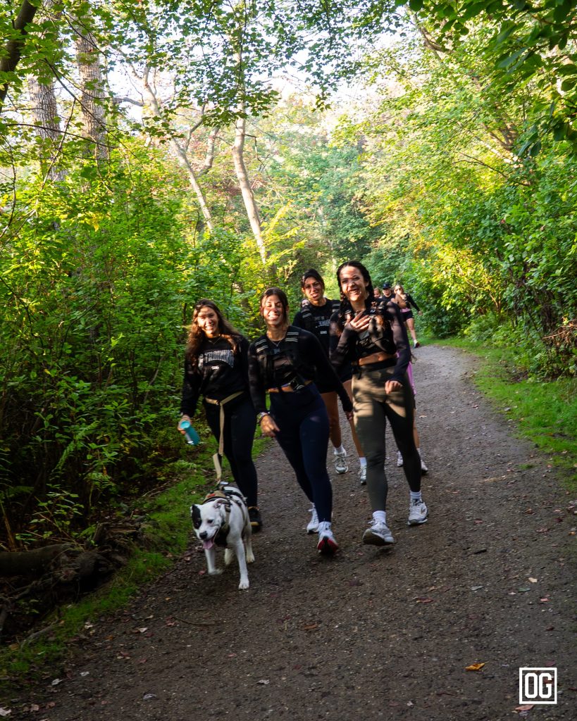Four OG Run Club members running together toward the camera along a wooded trail at Southards Pond Park, with a dog alongside them