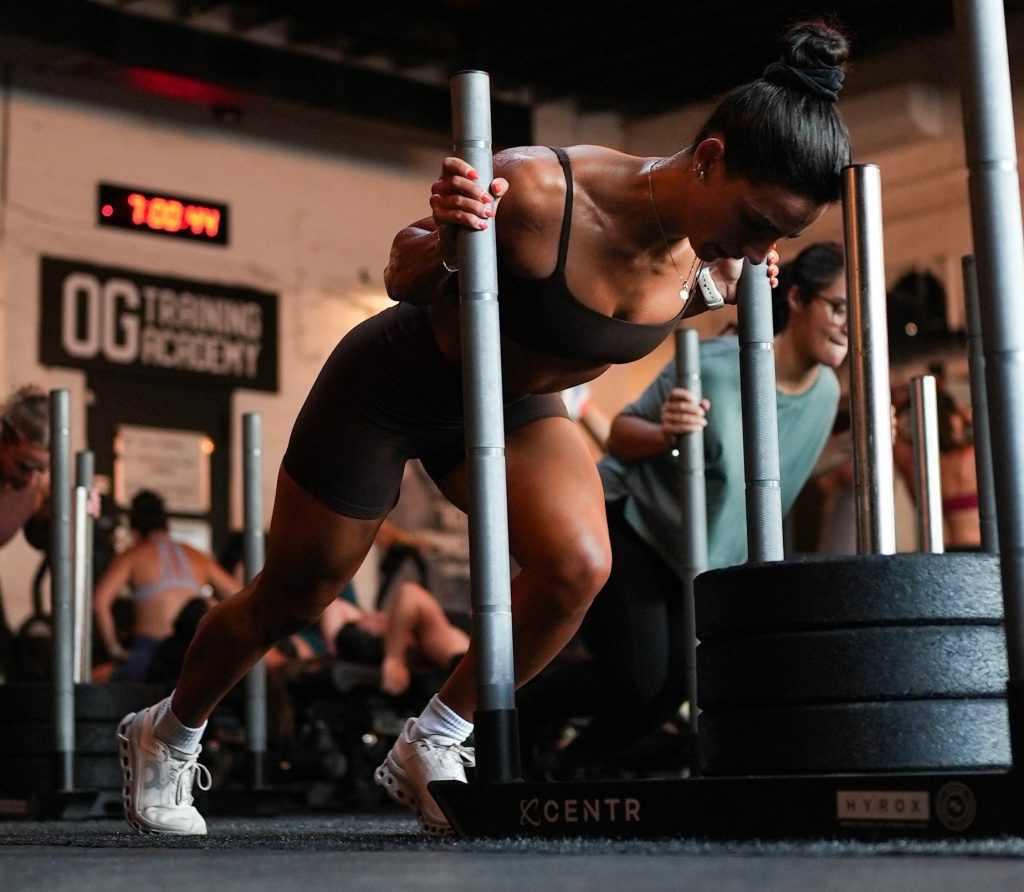 An athlete pushing a loaded sled on the training floor at OG Training Academy, with other members training behind her under the gym sign