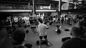 Wide view of a jiu-jitsu academy with students seated in a circle around an instructor at a We Defy Foundation event