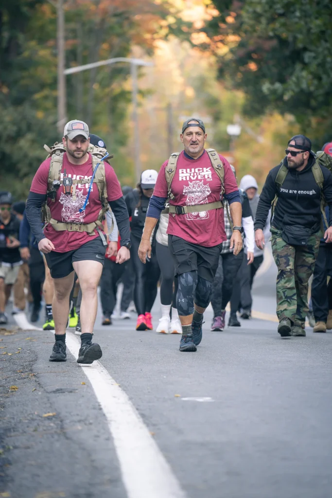 Ruckers in Strides For Service shirts walking a tree-lined road with loaded packs at Ruck The River
