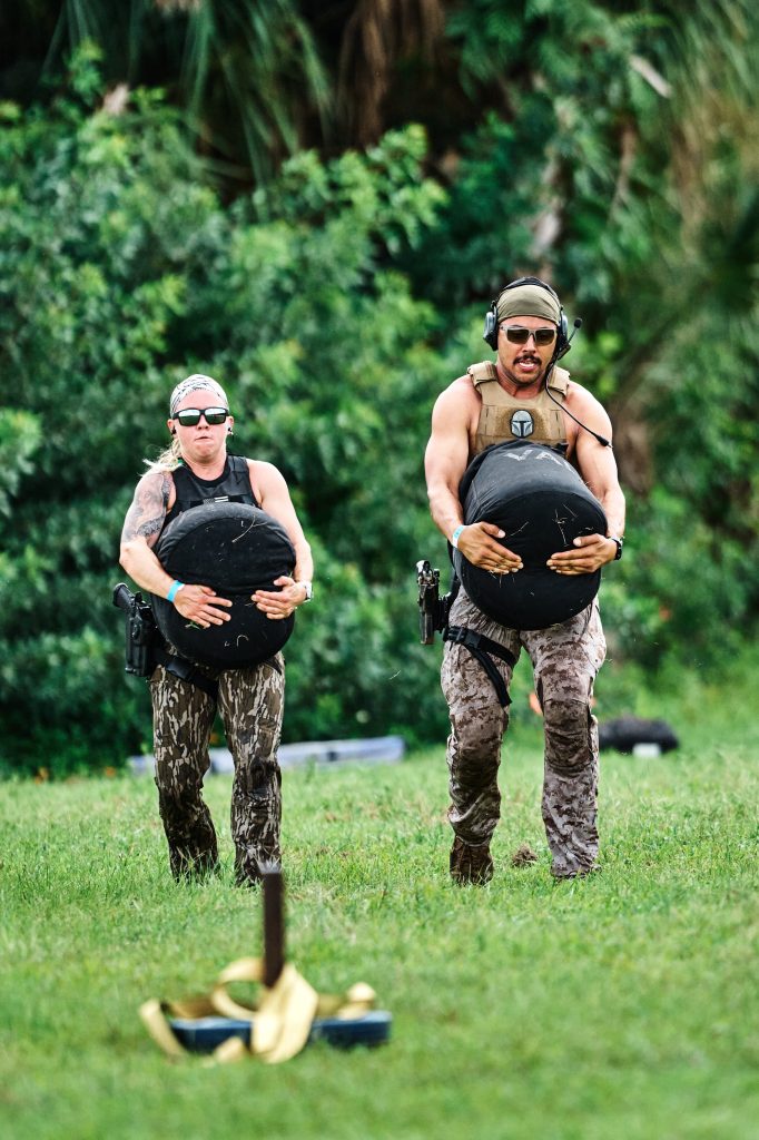 Two Patriot Games competitors in plate carriers and holstered pistols carrying heavy sandbags across grass during a timed stage