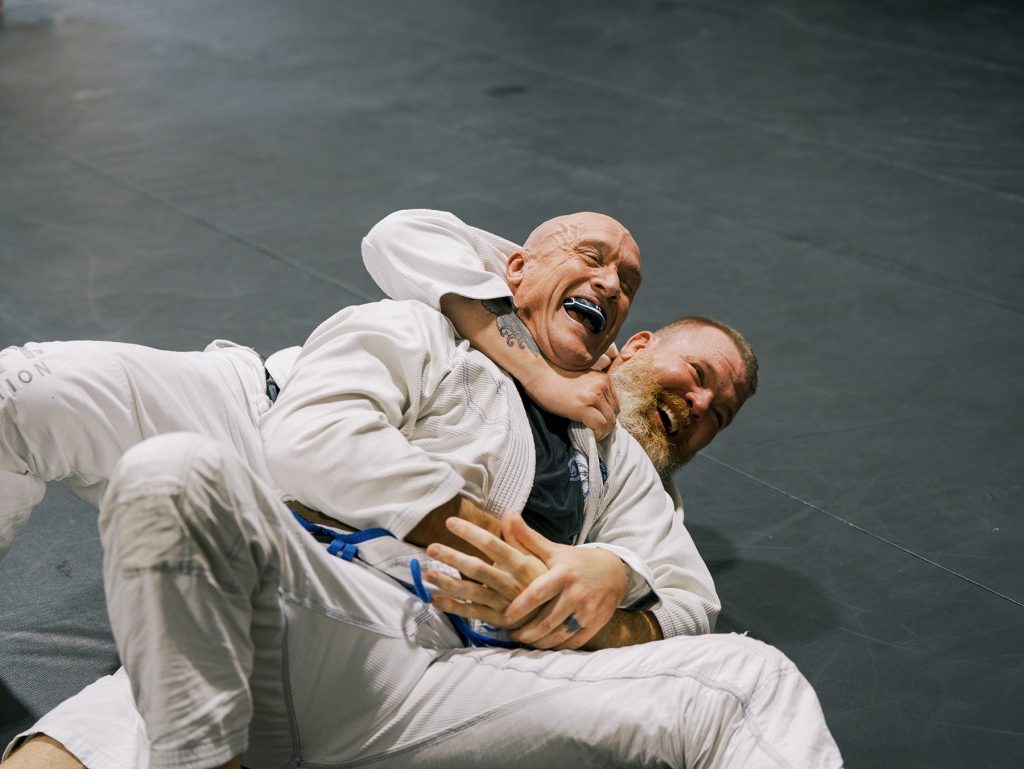 Two men in jiu-jitsu gis grappling on the mat and laughing at a We Defy Foundation event